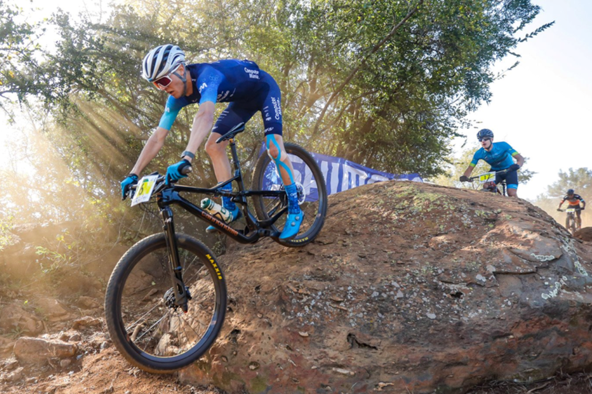MTB cross-country riders descending a rock feature in dappled forest light during the national series.