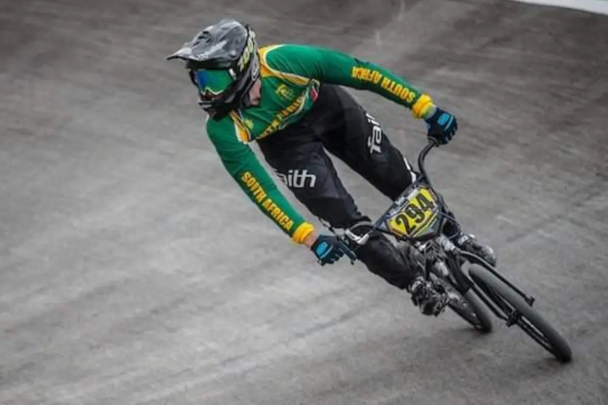 BMX rider in South Africa leaning into a berm on a grey track, green-and-gold jersey, action shot with race number visible.