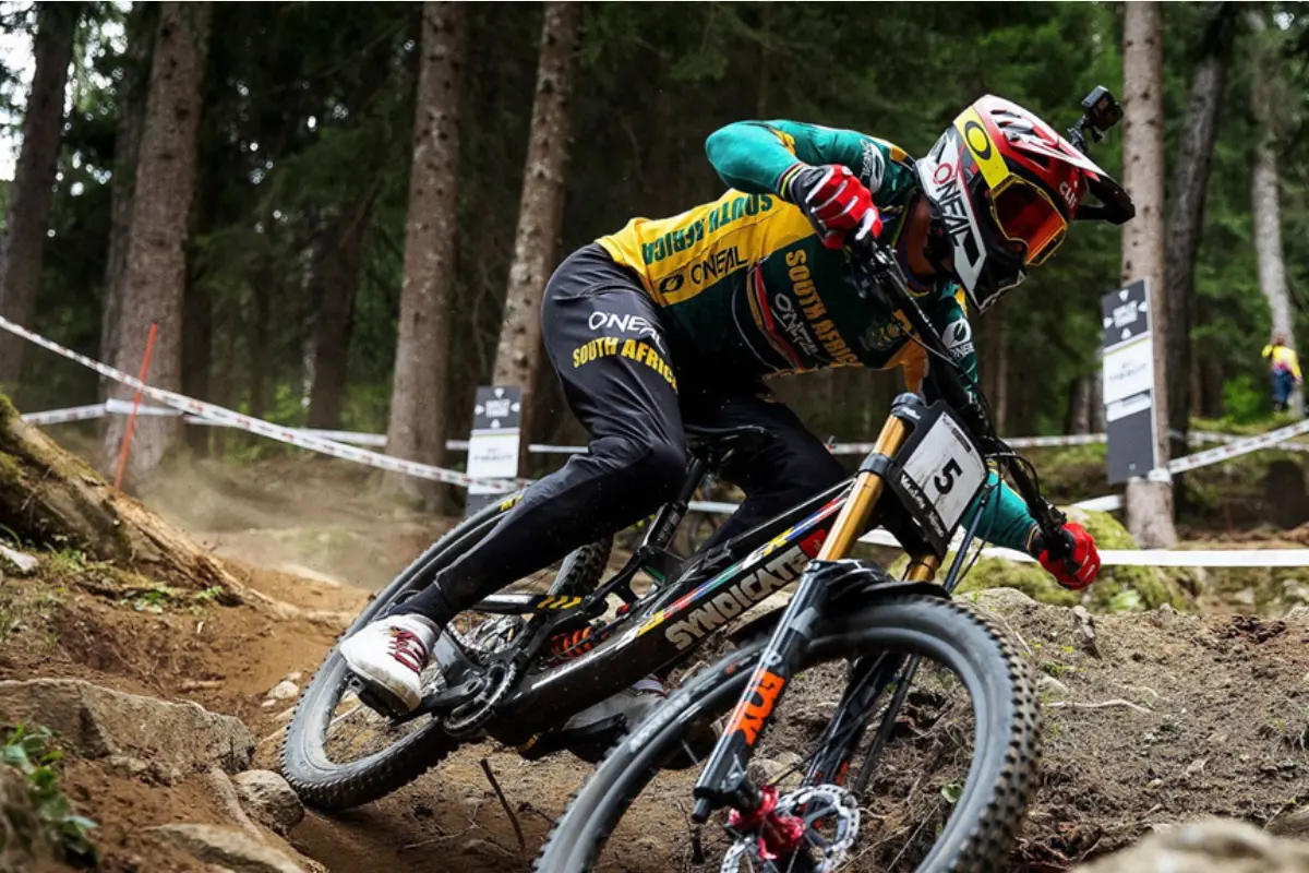 South African downhill rider in green-and-gold kit leaning through a forest berm, race tape and trees in background, bike no. 5.
