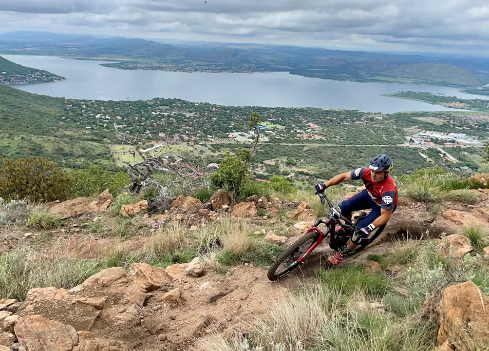 Enduro mountain biker leaning into a rocky corner on a steep trail with a lake and town in the background.