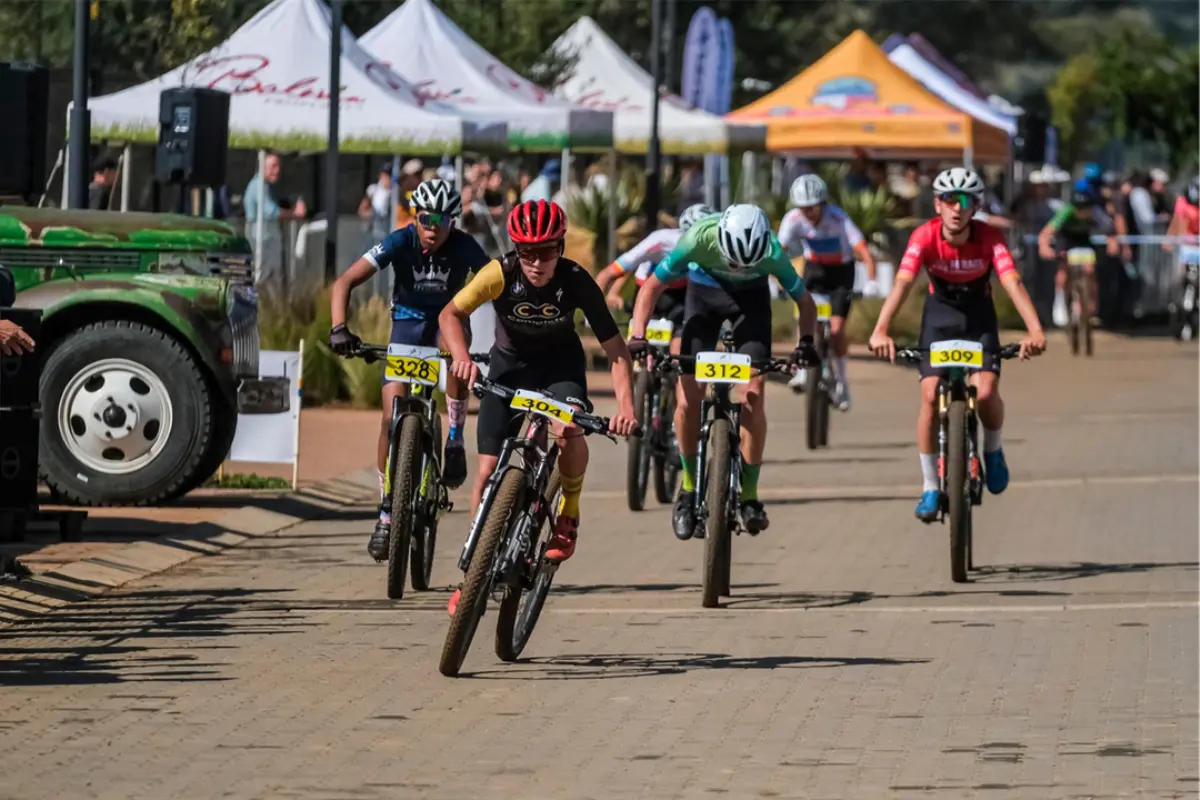 Junior MTB racers sprinting on a paved start/finish zone, front rider from Gauteng leading with number 304, tents and spectators in background.
