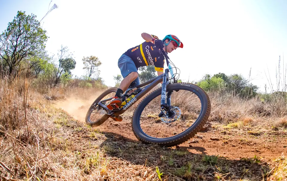 Enduro mountain biker in red helmet cornering hard on a dusty singletrack with dry grass and trees in the background.