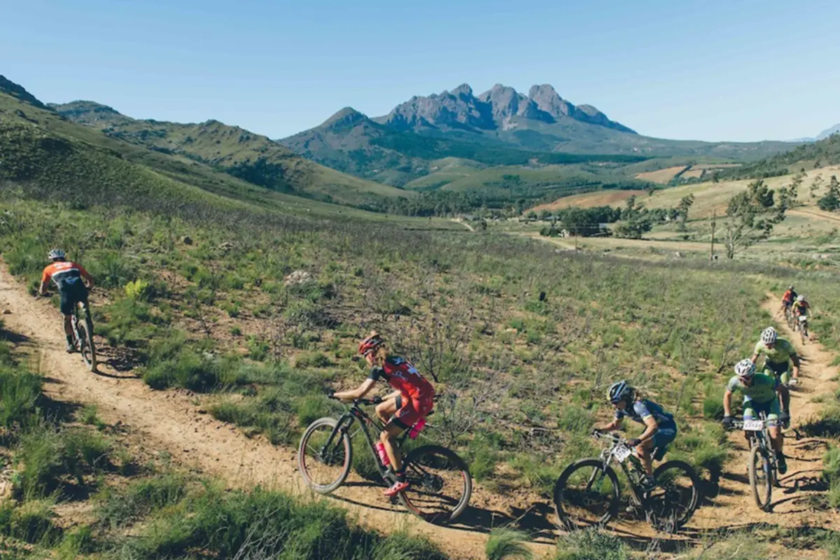 Mountain bikers racing along a dusty singletrack through open hills with a dramatic mountain range in the background.