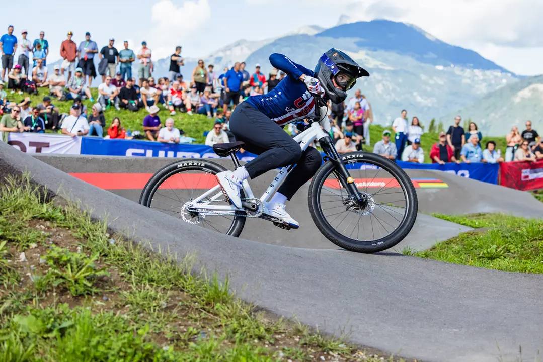 Mountain biker in full-face helmet racing around a paved pump track berm with spectators and mountains in the background.