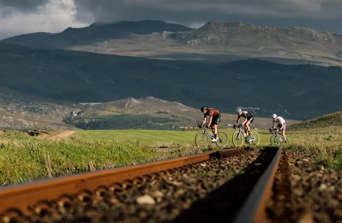 Three gravel cyclists riding alongside railway tracks with dramatic mountains in the background.
